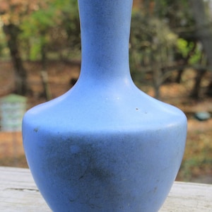 May include: A small, light blue ceramic vase with a rounded body and a narrow neck. The vase has a matte finish and a slightly textured surface.