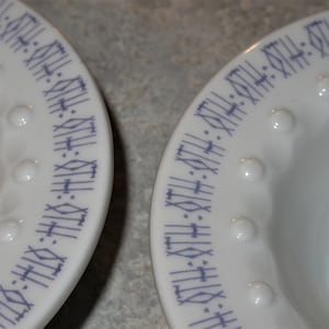 May include: Two white ceramic bowls with a purple geometric pattern around the rim. The bowls have a raised, textured rim.