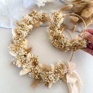 Può includere: Due cerchi circolari in metallo dorato decorati con fiori secchi bianchi e beige, tra cui gipsofila e code di lepre. Il cerchio più grande presenta un nastro beige chiaro legato sul fondo, mentre il cerchio più piccolo è tenuto in mano.