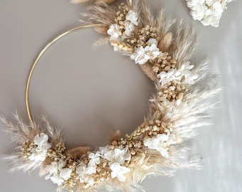 May include: A gold metal hoop wreath adorned with dried pampas grass, wheat, and white flowers. The wreath is a circular shape and is decorated with natural elements, creating a rustic and elegant aesthetic.