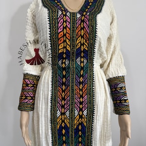 Puede incluir: Un vestido blanco bordado con coloridos patrones geométricos. El vestido tiene mangas largas y un escote en V. El bordado es intrincado y detallado, y los colores son vibrantes. El vestido es una prenda tradicional etíope.