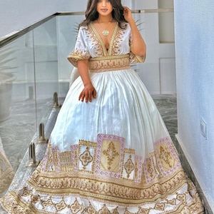 Puede incluir: Un vestido formal blanco con detalles bordados en oro y rosa. El vestido presenta un escote en V profundo, mangas cortas y una falda amplia. La falda tiene un diseño de borde detallado en oro y rosa. El vestido es usado por una persona.