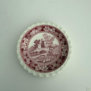 May include: A small, white ceramic saucer with a pink floral design. The design features a bridge, a building, and trees. The saucer has a scalloped edge.