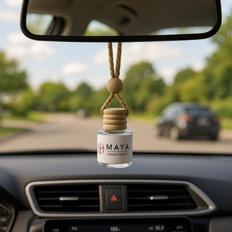 May include: A car air freshener with a glass bottle and wooden detailing, suspended from a rearview mirror. The label on the bottle reads "MAYA FRESH PEACH". The background shows a road and trees.