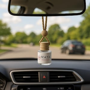 May include: A car air freshener with a glass bottle and wooden details hangs from a rearview mirror. The bottle is labelled "MAYA FRESH PEACH". The background shows a road and a car.
