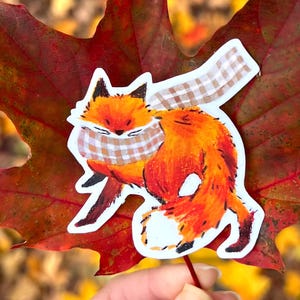 May include: A sticker featuring two orange foxes with a plaid scarf, placed on a red and green leaf. The foxes are depicted in a playful embrace. The background is blurred, showing autumnal colours and foliage.
