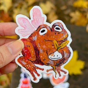 May include: A sticker depicting a cartoon frog with pink fairy wings, holding a small plant. The frog is orange with white eyes and a smiling expression. The sticker has a glittery effect.