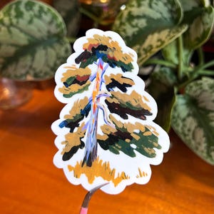 May include: A die-cut sticker with a painted tree design. The tree has a blue and white trunk, with green, brown, and yellow foliage. The sticker is held by pink tweezers against a blurred background of green leaves.