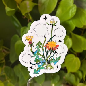 Könnte beinhalten: Ein weißer Aufkleber mit einer skurrilen Illustration von Löwenzahnpflanzen. Das Design umfasst gelbe und orangefarbene Blüten, grüne Blätter und einen kleinen Frosch. Schwarze Umrisse und Sterne.