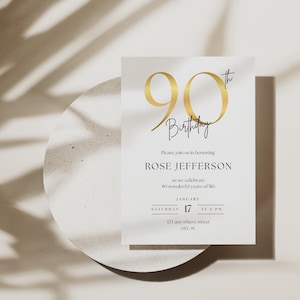 Op de afbeelding: Een witte uitnodigingskaart met gouden tekst en een gouden '90th' afbeelding. De kaart luidt 'Verjaardag' en nodigt gasten uit om de 90e verjaardag van Rose Jefferson op 17 januari om 18.00 uur te vieren. De kaart ligt op een wit, getextureerd oppervlak.