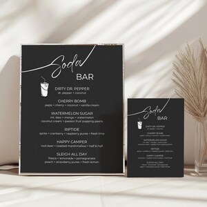 May include: Two black chalkboard signs with white text listing a menu of soda bar drinks. The signs are titled "Soda Bar" and list drinks like "Dirty Dr. Pepper", "Cherry Bomb", "Watermelon Sugar", "Riptide", "Happy Camper", and "Sleigh All Day".