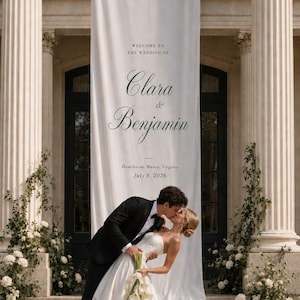 Könnte beinhalten: Ein hohes Hochzeitsbanner aus weißem Stoff mit dem Text "WELCOME TO THE WEDDING OF Clara & Benjamin Hawthorne Manor, Virginia July 8, 2026" in dunkelgrüner Schrift, das vertikal zwischen zwei großen Steinsäulen angebracht ist.