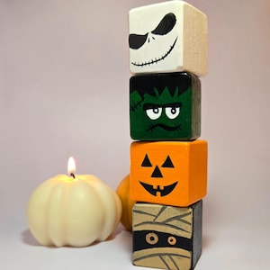 May include: A stack of four wooden blocks with painted Halloween character faces. The top block is white with a black Jack Skellington face, the second block is green with a Frankenstein face, the third block is orange with a pumpkin face, and the bottom block is brown with a mummy face.