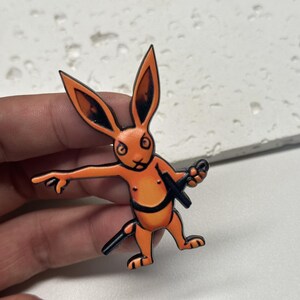 Puede incluir: Pin de esmalte naranja y negro de un conejo sosteniendo una espada. El conejo tiene una expresi&oacute;n amenazante y lleva un cintur&oacute;n negro.