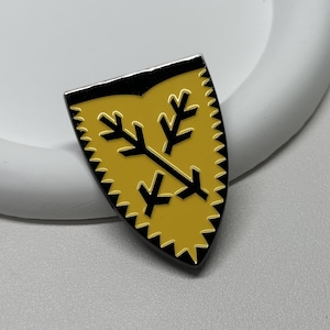 Puede incluir: Un pin de esmalte amarillo con forma de escudo con borde negro y un diseño negro de flechas cruzadas con puntas de flecha. El escudo tiene un borde dentado.