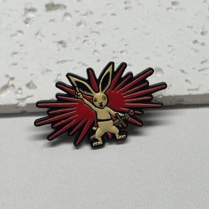Puede incluir: Un pin de esmalte negro y rojo con un conejo blanco sosteniendo una espada, de pie frente a una explosi&oacute;n roja y negra.