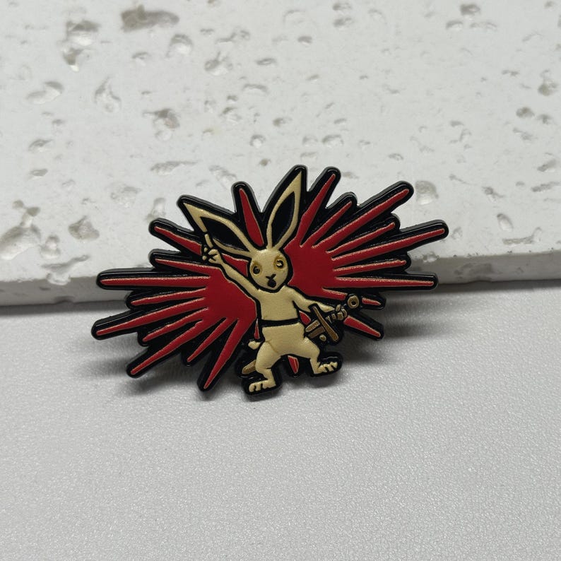 Puede incluir: Un pin de esmalte negro y rojo con un conejo blanco sosteniendo una espada, de pie frente a una explosi&oacute;n roja y negra.