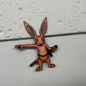 Puede incluir: Pin de esmalte naranja y marr&oacute;n de un conejo sosteniendo una espada. El conejo tiene una expresi&oacute;n seria y est&aacute; apuntando hacia la izquierda.