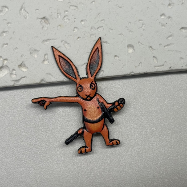 Puede incluir: Pin de esmalte naranja y marr&oacute;n de un conejo sosteniendo una espada. El conejo tiene una expresi&oacute;n seria y est&aacute; apuntando hacia la izquierda.