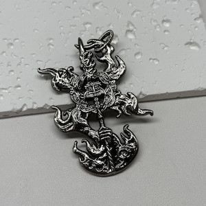 May include: A silver pendant with a detailed design of a creature with flames. The pendant is made of metal and has a textured surface.