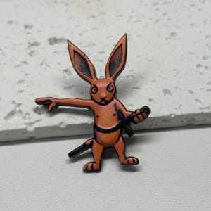 Puede incluir: Pin de esmalte naranja de un conejo sosteniendo una espada y apuntando con su mano izquierda. El conejo tiene una expresi&oacute;n seria en su rostro.