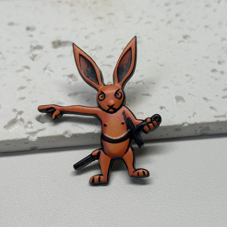 Puede incluir: Pin de esmalte naranja de un conejo sosteniendo una espada y apuntando con su mano izquierda. El conejo tiene una expresi&oacute;n seria en su rostro.