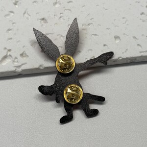 Puede incluir: Pin de esmalte negro de un conejo de dibujos animados con un dise&ntilde;o rojo y blanco. El pin tiene una parte posterior dorada con dos cierres.