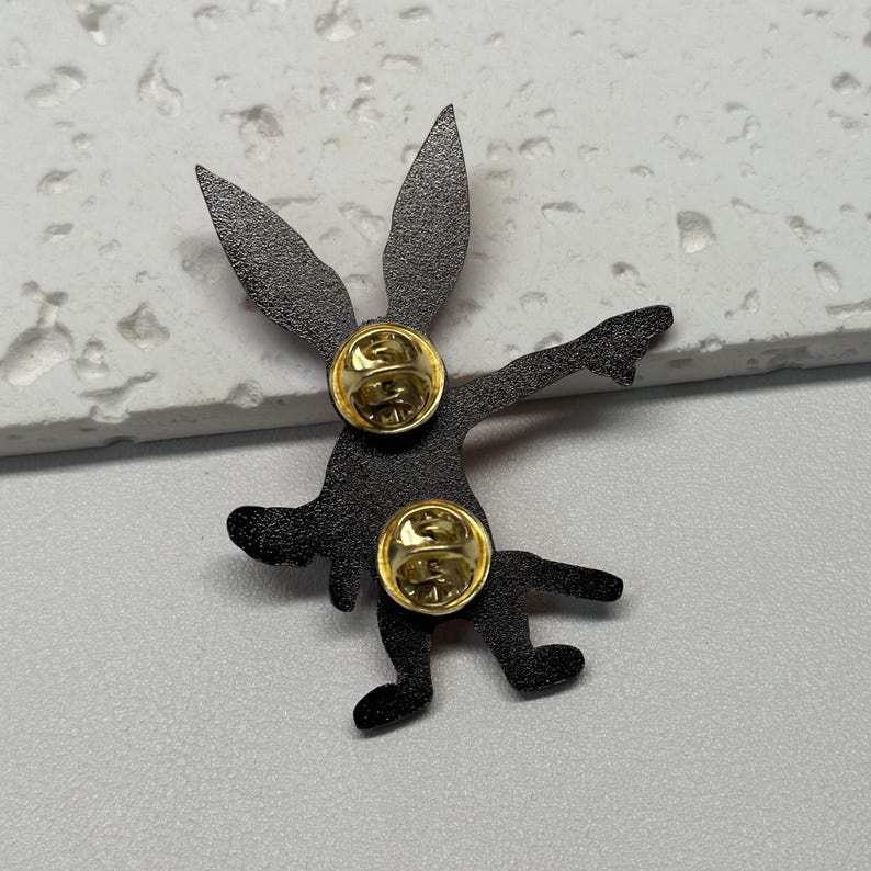 Puede incluir: Pin de esmalte negro de un conejo de dibujos animados con un dise&ntilde;o rojo y blanco. El pin tiene una parte posterior dorada con dos cierres.