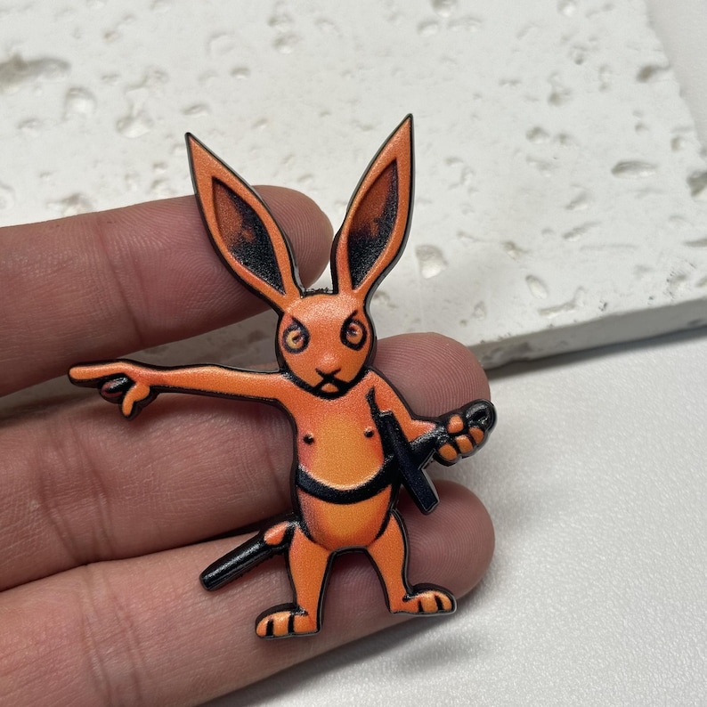 Puede incluir: Pin de esmalte naranja y negro de un conejo sosteniendo una espada y apuntando con su mano izquierda.