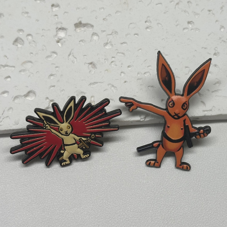Puede incluir: Dos pines de esmalte con conejos de dibujos animados. Un conejo es naranja y sostiene una espada. El otro conejo es blanco y est&aacute; rodeado de rayos rojos y blancos.