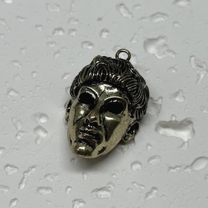 May include: A gold-toned pendant featuring a detailed sculpted face with a crown of braided hair. The pendant has a loop at the top for attaching a chain.
