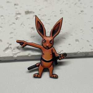 Puede incluir: Pin de esmalte naranja de un conejo con contornos negros. El conejo sostiene una espada y apunta con su pata derecha.