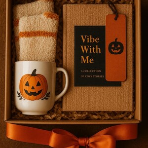 Op de afbeelding: Een Halloween-cadeaupakket met een witte mok met een pompoenontwerp, een boek met de titel "Vibe With Me", zachte sokken en een oranje lint. De doos is gevuld met decoratief stro.