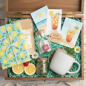 Op de afbeelding: Een houten kist gevuld met zomerse items. Bevat een ingepakt cadeau met een citroenpatroon, ansichtkaarten met ijsthee-illustraties, een witte mok en kleine glazen potjes. De kist is bekleed met groen papier en versierd met madeliefjes.