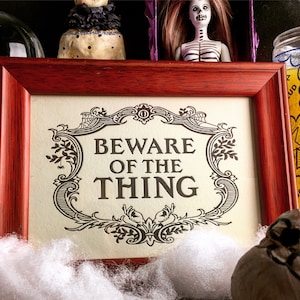 以下が含まれることがあります： クリーム色の背景に黒い文字で「Beware of the Thing」と書かれた、ヴィンテージデザインの額縁入り看板です。
