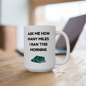 May include: White ceramic mug with the text "ASK ME HOW MANY MILES I RAN THIS MORNING" in black. Below the text is a graphic of teal running shoes. The mug is on a wooden surface.