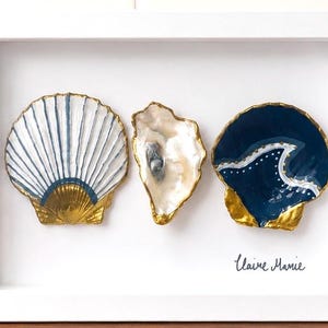 May include: A white rectangular frame containing three decorative seashells. The left shell has blue stripes and a gold base. The center shell is iridescent white. The right shell is dark blue with a white wave design and gold edges. Text reads Claire Marie.