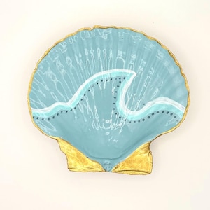 May include: A decorative scallop shell painted in a light blue hue with a gold-painted edge. The interior features a hand-painted white wave design accented with a row of small black dots across the center, creating a distinct visual contrast.