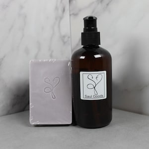 Può includere: Una bottiglia di vetro marrone con un dosatore a pompa nero accanto a una saponetta lilla chiaro. La bottiglia ha un'etichetta bianca con il testo "Saul Goods". Il sapone ha un design a forma di cuore.