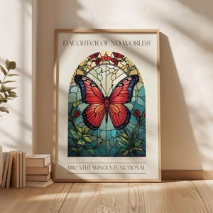 Puede incluir: Una impresión enmarcada con un diseño de vitral que presenta una mariposa roja con marcas negras sobre un fondo azul y verde. El texto "Daughter of No Worlds" y "Breathtakingly Functional" está impreso debajo de la imagen.
