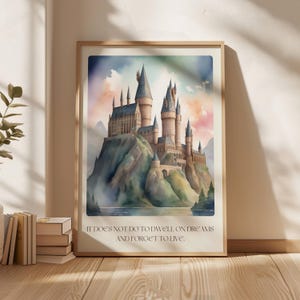 以下が含まれることがあります： 魔法使いと魔法使いのための架空の学校であるホグワーツ城の水彩画。丘の上にあり、ハリーポッターシリーズからの引用が書かれています。「夢にふけって生きることを忘れてはいけない。」