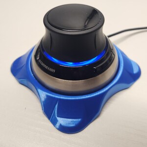 May include: A 3Dconnexion SpaceMouse, a black and silver device with a blue illuminated ring, sits on a blue base. The device has a sleek, modern design and is connected to a black cable.