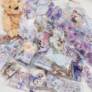 May include: A collection of decorative stickers with floral and fantasy designs. The stickers, packaged in clear plastic, feature roses, fairies, and other whimsical elements. A plush teddy bear is visible in the upper left corner.