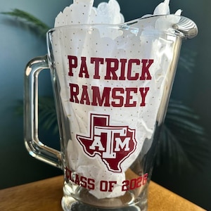 May include: A clear glass pitcher with a handle, featuring the name "PATRICK RAMSEY" in maroon block letters. Below the name is the Texas A&M logo and "CLASS OF 2026". The pitcher is filled with white tissue paper.