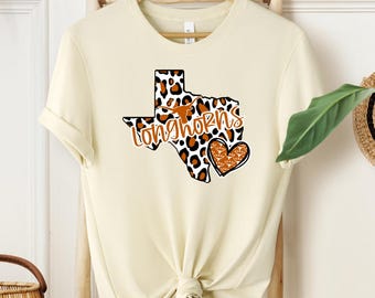 May include: A cream-colored t-shirt with a leopard print outline of the state of Texas, the word "Longhorns" and a heart with a leopard print design.