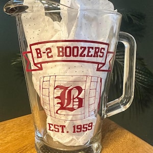 May include: A clear glass pitcher with a handle and a pouring spout. The front features a red graphic design with the text B-2 BOOZERS on a banner, a large letter B inside a barrel outline, and the text EST. 1959 printed below the barrel.