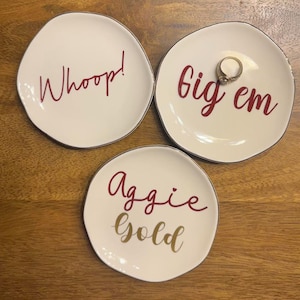 May include: Three white ceramic ring dishes with gold trim. The dishes have red lettering. One dish says "Whoop!" Another dish says "Big em". The third dish says "Aggie gold".