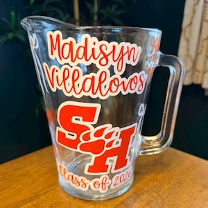 May include: Clear glass pitcher with a handle. The pitcher has red and white lettering that reads "Madisyn Villalovos" and "SA" with a paw print design. The text "Class of 2026" is also visible.