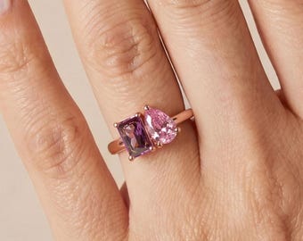 Könnte beinhalten: Ein roségoldener Ring mit zwei Edelsteinen: einem rechteckigen, dunkelvioletten Stein und einem birnenförmigen, rosa Stein. Der Ring wird auf einer Hand mit polierten Nägeln präsentiert.