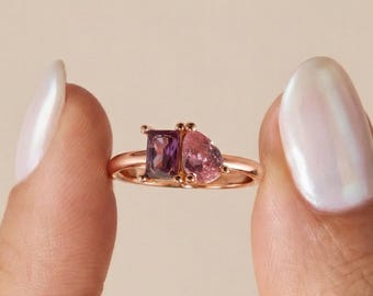 Könnte beinhalten: Ein Ring aus Roségold mit zwei nebeneinander liegenden Edelsteinen: einem rechteckigen violetten Stein und einem birnenförmigen rosa Stein. Das Band ist dünn und poliert und unterstreicht die kontrastierenden Formen und Farben der Steine.